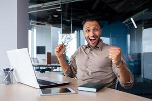 Successful Indian programmer celebrating successful completion of testing new software, man looking at camera, satisfied with achievement results, working with laptop, inside office, online winner. photo