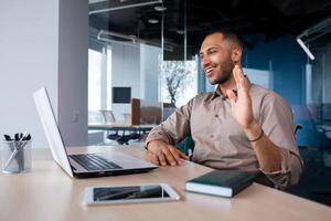 Happy successful and satisfied man talking on call using laptop inside office, businessman waving hand smiling and greeting colleagues and partners, remote online meeting presentation. photo