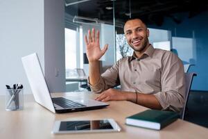 Portrait Satisfied African American businessman in office, working on laptop, tapping on keyboard, greeting waving hand looking at camera. photo