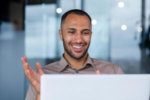 Close-up of a happy successful hispanic businessman making a call from a laptop in a modern office, a young programmer is happy about a successful job, smiling at the computer screen. photo