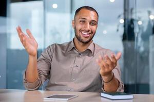 Attractive hispanic businessman having call in office, gesturing with hands, smiling, talking to clients, webcam view. photo