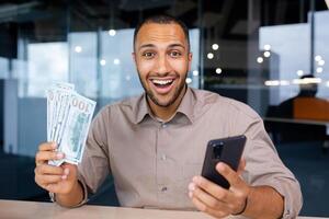 Excited man sitting in modern office holding smartphone and wad of dollars in hands, happy with winning, smiling and looking at camera. photo
