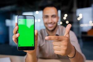 Smiling man holding a phone with a green screen in his hands, showing it to the camera and pointing at it with his index finger, looking at the camera. photo