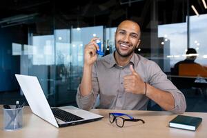 African american guy sitting in office with asthma inhaler in hand, showing thumbs up like, smiling and looking at camera. photo