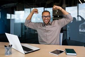 Happy man working in office showing muscles raising arms up, happy at workplace, smiling and looking at camera. photo