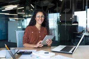 Happy and successful hispanic woman working inside modern office building, business woman using tablet computer smiling and looking at camera worker doing paperwork. photo
