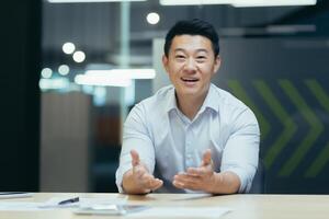 Online webinar. Portrait of an active young Asian businessman. He conducts an online webinar on camera, sits at a table in a modern office, talks, explains, gestures with his hands, smiles. photo