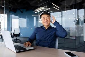 Portrait of happy asian worker in modern office, man looking at camera and smiling, boss programmer in shirt using headphones to watch and listen to music at work. photo