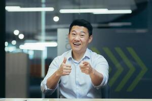 Portrait of a successful and happy Asian male businessman. He sits in the office at the work table, shows with his hands to the camera winnings, good luck, motivates, says you can do it. photo