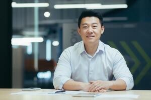 Portrait of a young Asian freelancer, designer, architect. He sits at the desk in the office, holds the phone, works with documents, looks into the camera, smilling. photo