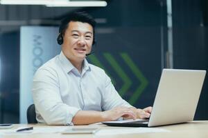 Portrait of successful asian businessman man with call headset looking at camera and smiling, tech support consultant working in office with laptop photo
