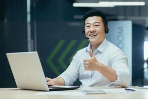 A young male student sits in an office, co-working space, library at a desk in a headset. learns online remotely from a laptop. He looks at the camera, points a super finger, smiles photo