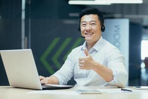 Successful asian businessman hardworking support tech with headset for call looking at camera and smiling showing thumbs up, man working in office with laptop photo