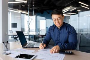 Successful and happy investor working with documents and charts inside office, Asian businessman behind paper work calculates bills with calculator, man at work with laptop happy with achievements. photo