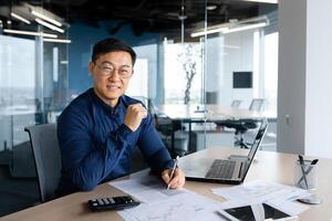 Portrait successful satisfied asian financier accountant, man in shirt and glasses working inside office using calculator and laptop for accounts and financial documents reports, looking at camera. photo