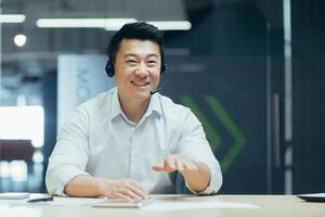 Online business training. Young smiling Asian man sit and talk in the office in front of the camera in a headset, conducts an online meeting, webinar photo