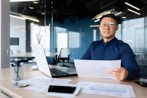 Portrait of a young Asian architect, designer, engineer. Sitting at the desk in the office, holding papers. Works with documents, projects, plans. He looks at the camera, smiles. photo