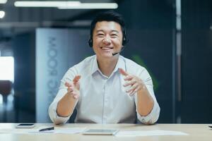 Asian man with headset talking on call, looking into web camera, smiling cheerfully and gesticulating with hands, tech support photo