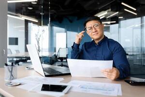 Serious young Asian architect, designer, engineer. Sitting at the desk in the office, holding documents and a pencil. The problem is working with projects, plans. Looking worriedly at the camera. photo
