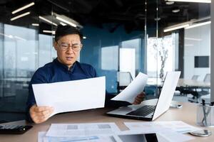 Asian businessman not satisfied with achievement results, boss looking at documents and upset, man in glasses and shirt working inside office with paperwork using laptop. photo