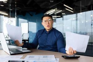 Problems at work. Worried young Asian man, businessman, freelancer. He sits in the office at a table with a laptop, holds documents in his hands. Spreads his hands, looks at the camera confused. photo