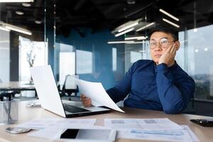 Serious thinking asian man working with documents inside office, concentrated businessman with contracts and bills reading and calculating reports, mature financier with documents. photo