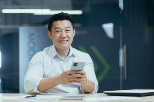 Happy Asian man using phone in office, businessman smiling and looking at camera typing message and browsing internet page photo
