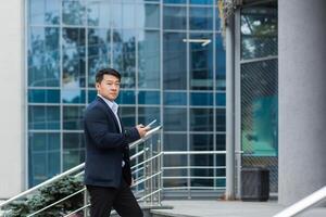 A young handsome Asian man goes to a meeting, an interview for a job in an office center. He is standing on the stairs with a phone and documents in his hands, looking at the camera. photo