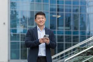 Portrait of Asian businessman outside modern office building, boss in business suit using phone smiling and looking at camera, man typing message and browsing web pages online. photo