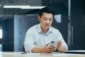 A break at work. A young Asian businessman is using the phone at his desk in a modern office. He holds it in his hands, types a message, makes a phone call. photo