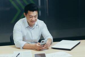 Asian man using phone in modern office, typing message and reading news while sitting at desk, businessman at work photo