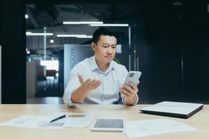 Confused Asian guy holding smart phone feels concerned thinking over received message. Mobile phone everyday usage, unpleasant news, waiting for important call, low signal device problems concept photo
