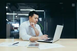 Serious and thoughtful young man of Asian origin programmer, developer works in office at desktop at laptop. photo