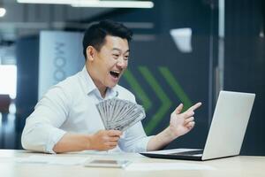 A happy and smiling young Asian businessman concluded a profitable financial deal with clients and partners. He sits in the office at a table with a laptop, shows and holds g of money in his hands. photo