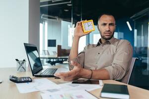 Dissatisfied hispanic man looking at camera and showing clock, businessman frustrated waiting for colleague to be late, working inside office with documents using laptop inside office. photo