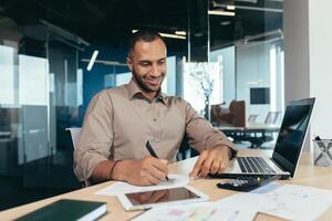 Successful african american businessman inside office doing paperwork, man writing information, worker using laptop for work sitting with bills and contracts smiling and satisfied with result. photo