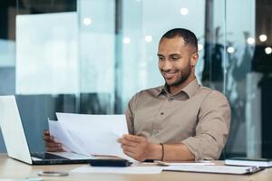 Successful businessman inside the office reading a financial report, hispanic man holding a contract in his hands and smiling with satisfaction, good achievement results, working on a laptop. photo
