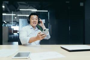 A young Asian man in headphones sits at the office table, holds the phone in his hands, looks at the screen, selects a playlist, a podcast. listening to music, relaxed, break and rest at work, study. photo