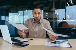 Portrait of disappointed young hispanic financier, businessman looking at camera in despair and holding failed financial report of business in hands, man working inside office building paperwork. photo