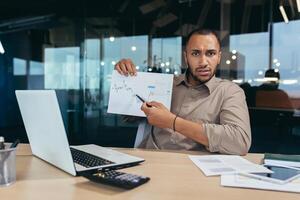 Portrait disappointed businessman inside office, african american man showing document graph with bad financial performance to camera, financier in overcoat working inside office using laptop. photo