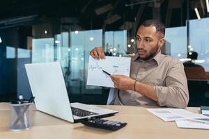 Dissatisfied businessman shows a graph with bad achievements to the camera on a call, a Latin American man reports negative results at an online meeting of company managers, a man at work. photo