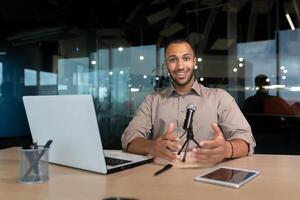 Portrait of a successful African American influencer, man smiling and looking at the camera, using a laptop and a professional microphone at work to record podcasts and read audio books online. photo