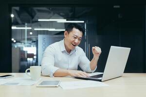 A young Asian man sits in the office at the table, rejoices at the victory and the win, looks at the laptop, participates in the auction, places bets. He smiles, shows a yes gesture with his hand photo