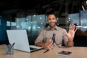 Successful hispanic businessman smiling and looking at camera, portrait of satisfied man in middle of office using microphone and laptop for call, waving hand in greeting gesture. photo
