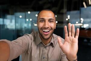 Smiling and cheerful businessman in office taking selfie photo on phone and talking on call with colleagues and friends using smartphone, african american man waving at camera greeting gesture.