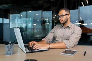 Serious and concentrated hispanic programmer working inside office, man in shirt writing codes for software using laptop. photo