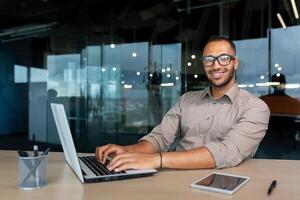 Portrait of successful hispanic businessman inside office, man with laptop working typing on keyboard smiling and looking at camera. photo