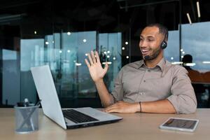 Online training. A young African-American male student sits in an office at a desk in front of a laptop in a headset. Learns through a call, says hello, smiles. photo