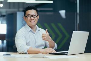 Portrait of successful asian businessman in office, man working with laptop typing smiling and looking at camera holding finger up, gesture of affirmation photo
