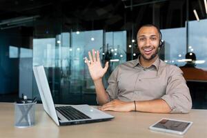 Portrait of tech support and online customer service worker, man with call headset smiling and looking at camera, african american man in shirt working inside office using laptop. photo